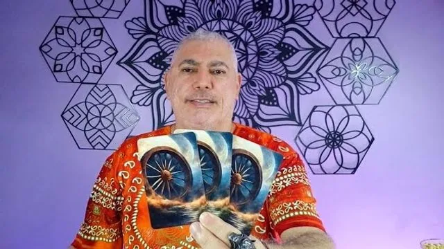 Henrique T’Oyá de camisa laranja estampada, com três cartas azuis com uma roda, diante da parede de mandalas