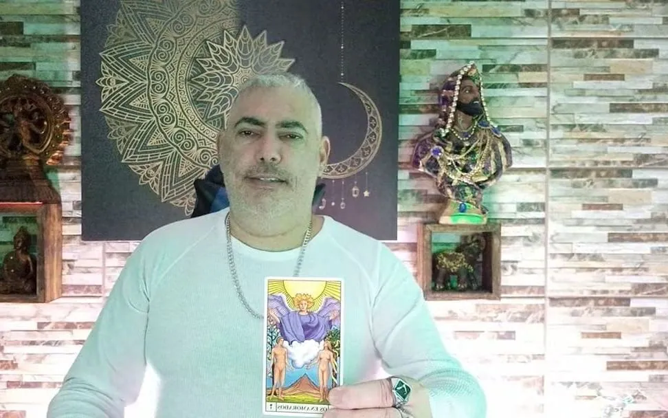 Henrique T’Oyá no estúdio dos vídeos, com a mandala dourada e a lua atrás, mostrando uma carta do tarô