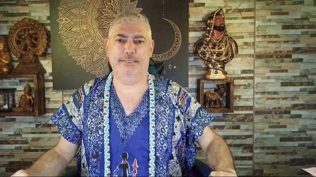 Henrique T’Oyá no estúdio dos vídeos, de bata azul e guias, com a mandala dourada e a lua atrás