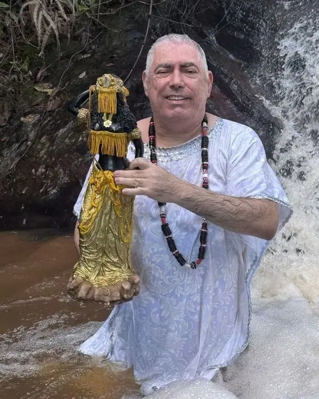 Henrique T’Oyá na cachoeira, de branco e com guias, segurando uma imagem dourada